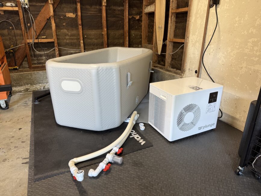 An inflatable ice bath tub next to a white plunge chiller unit, connected by hoses, set up in a garage with exposed wall studs.