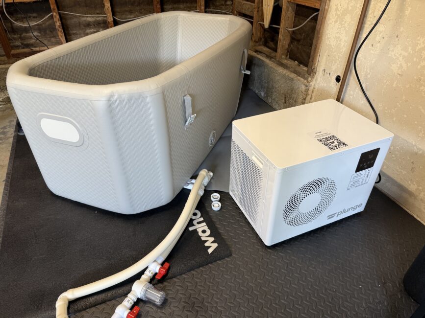 Inflatable ice bath tub connected by hoses to a white water chiller unit, set up on black mats in a partially finished room.