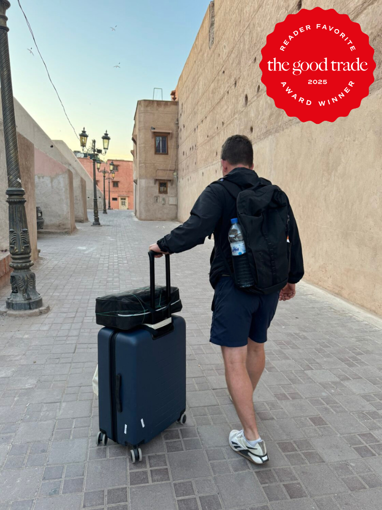 A man walks down a cobblestone street with a suitcase and backpack; a “The Good Trade 2025 Reader Favorite Award Winner” badge is in the top right corner.