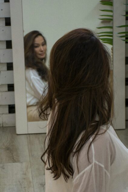 A woman with long brown hair, wearing a white shirt, sits on a wooden floor facing a mirror that reflects her face.