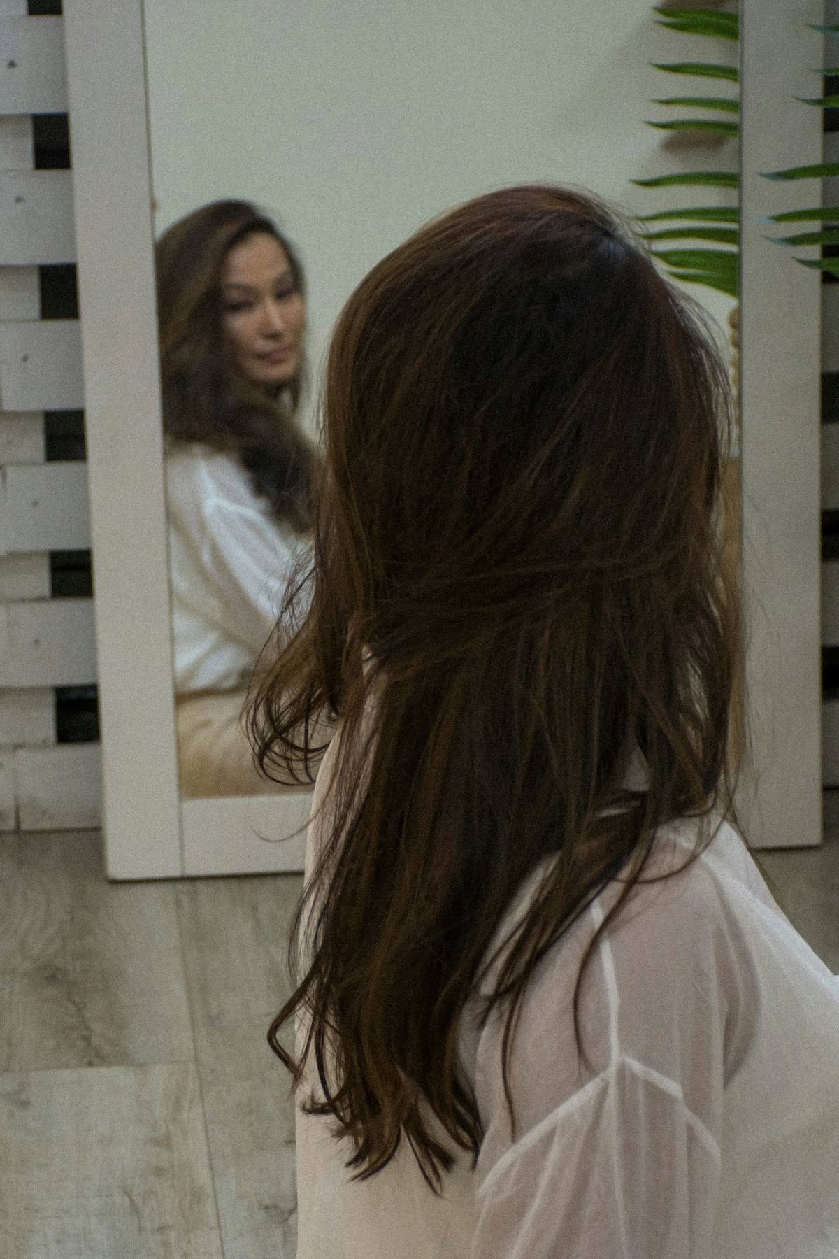 A woman with long brown hair, wearing a white shirt, sits on a wooden floor facing a mirror that reflects her face.