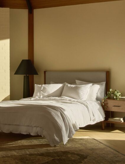 A neatly made bed with white linens and multiple pillows sits beside a wooden nightstand with a vase of flowers, under soft natural light in a minimalist bedroom.