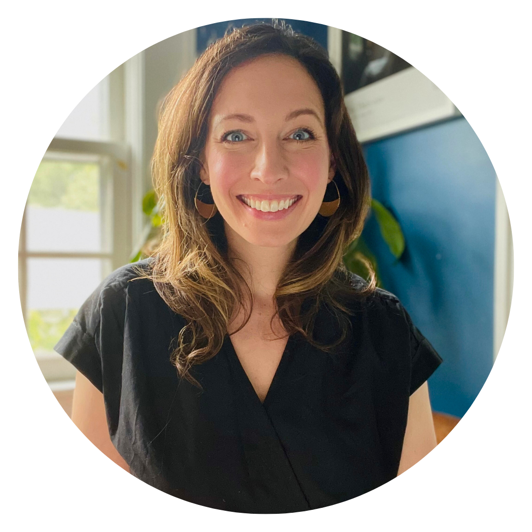 Woman with long brown hair wearing a black top stands indoors, smiling at the camera. Blue wall and window with greenery in the background.