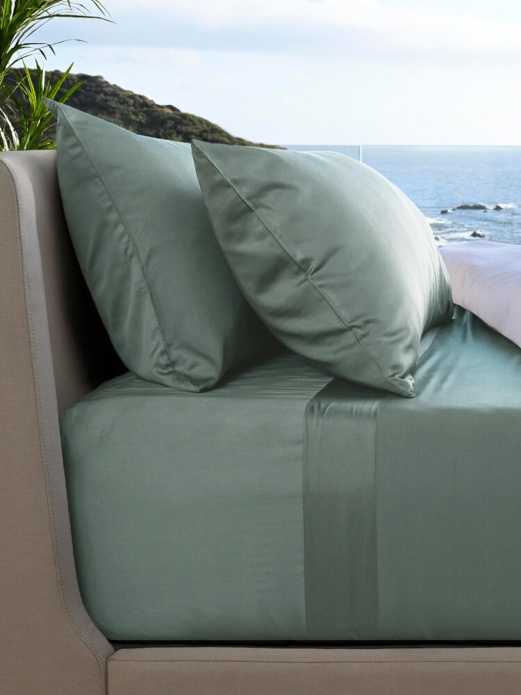 A neatly made bed with green sheets and pillows is positioned next to a window overlooking the ocean and a distant coastline.