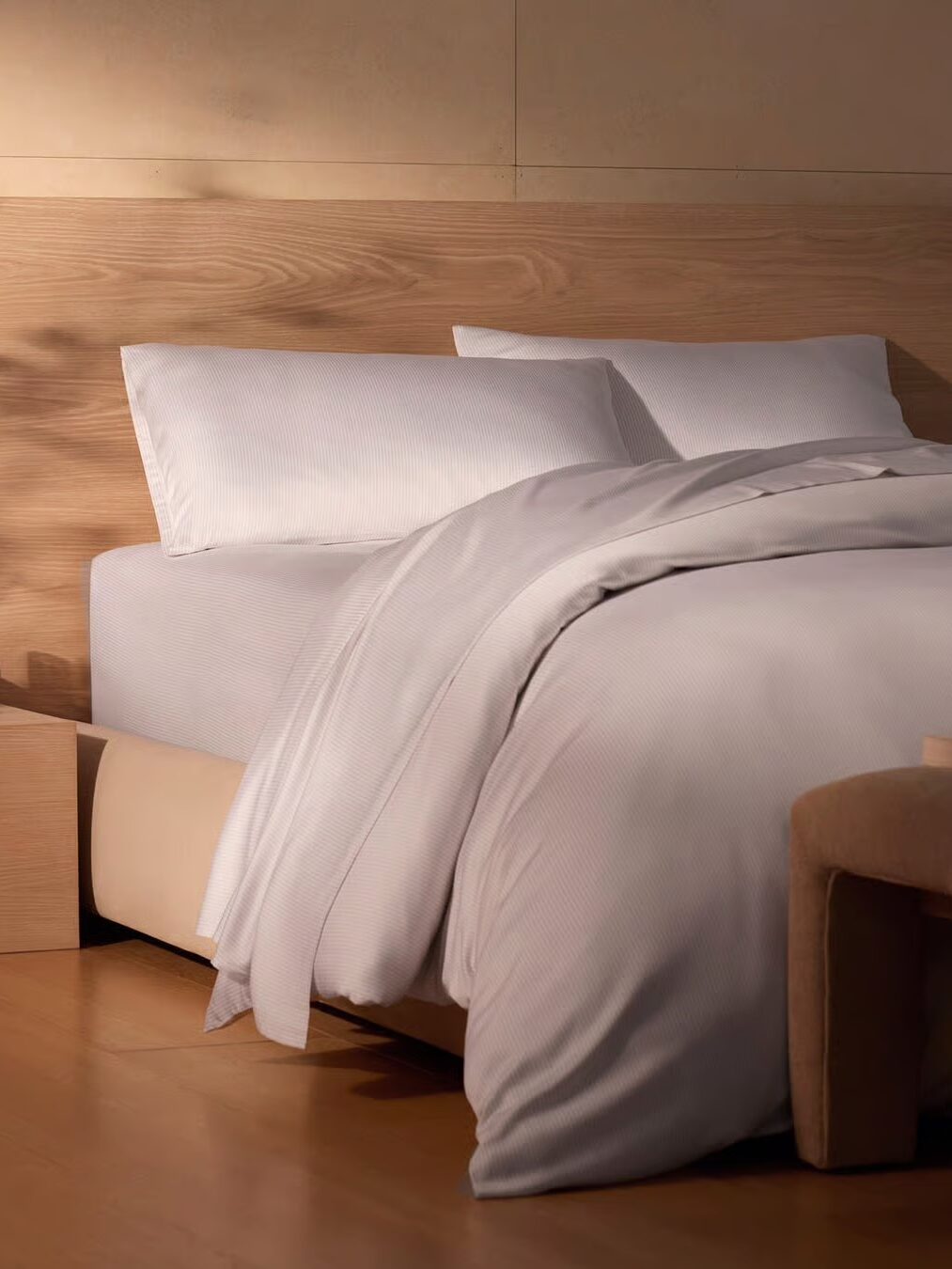 A neatly made bed with white linens and two pillows, set against a wooden headboard in a minimalist bedroom with light wood flooring.
