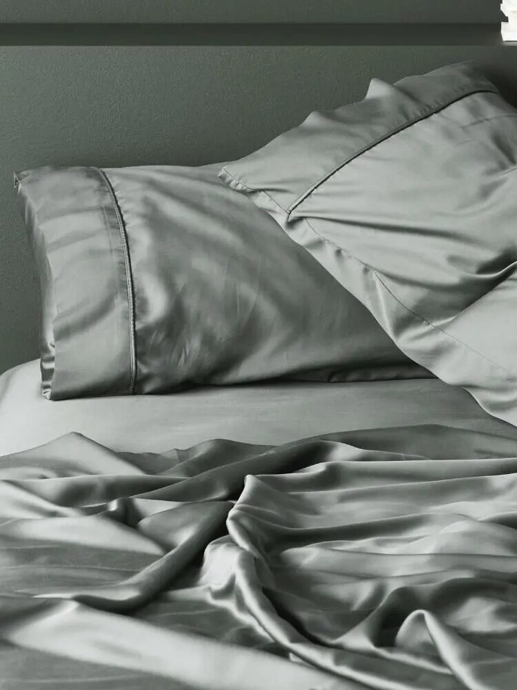 A neatly made bed with grey satin pillowcases and sheets, slightly rumpled, against a dark grey wall.