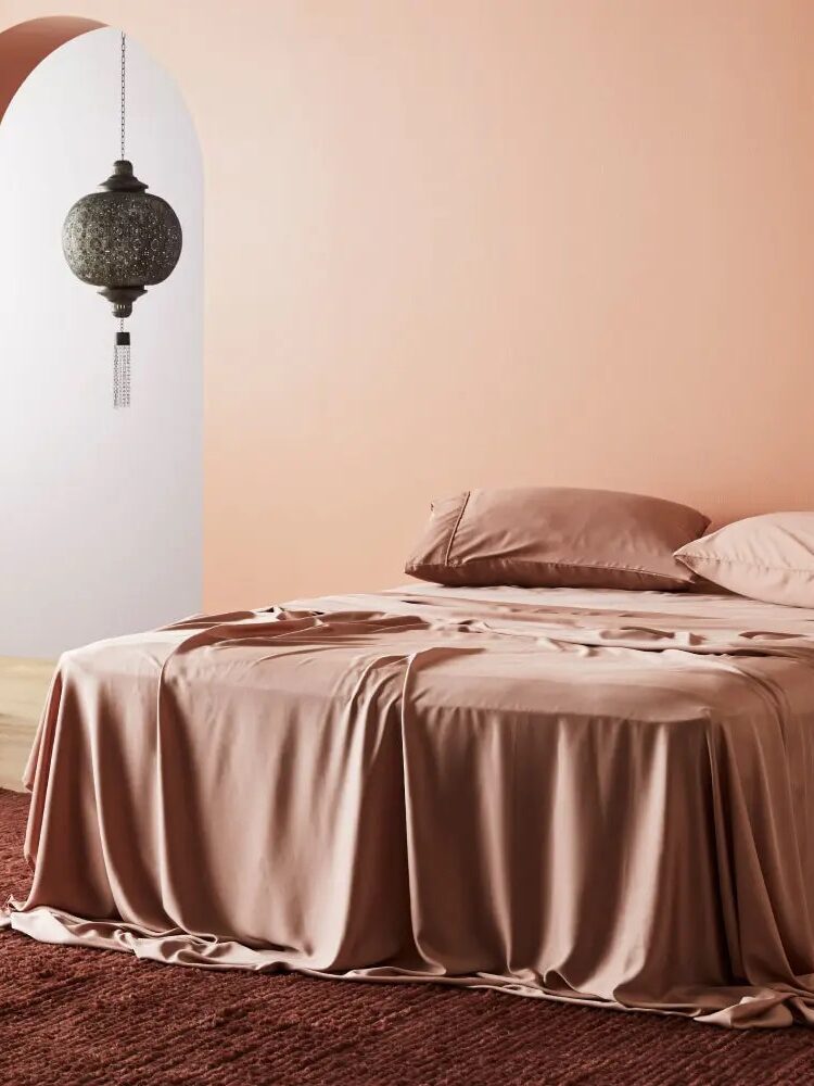 A neatly made bed with a draped dusty pink bedspread and pillows stands on a matching rug. An arched doorway and a hanging ornate lantern are visible in the background.