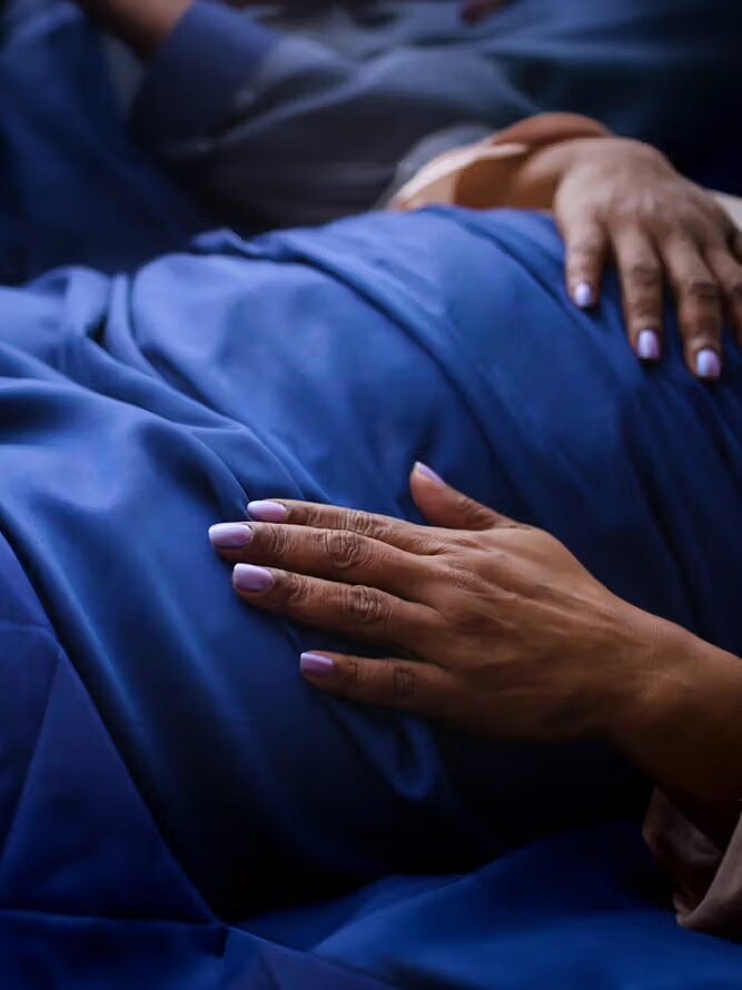 A close-up of hands resting on a person covered with a blue blanket, with purple-painted fingernails visible.
