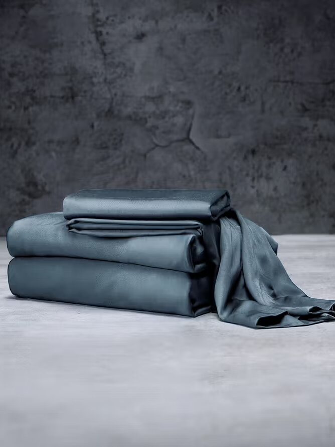 A neatly folded set of dark gray bed sheets on a light surface with a textured gray wall in the background.