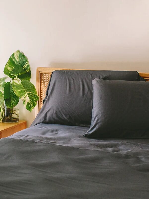 A neatly made bed with dark gray sheets and pillows beside a small wooden nightstand and several green potted plants against a white wall.