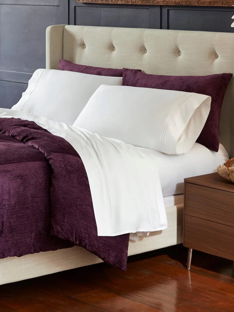 A neatly made bed with white sheets and pillowcases, a dark purple comforter and shams, set against a beige upholstered headboard in a modern bedroom.
