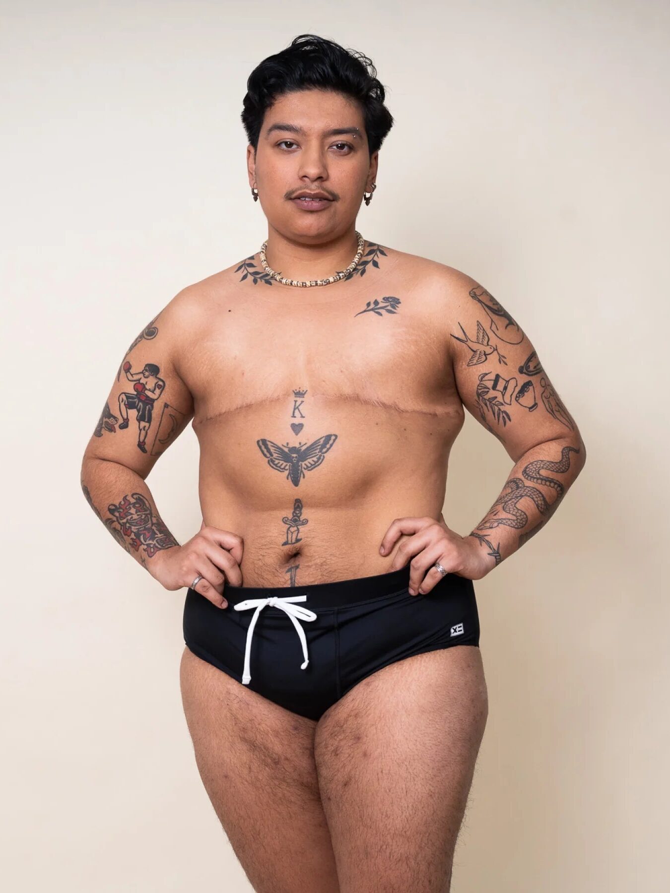 A person with short dark hair and chest scars stands against a plain background, wearing black swim briefs and displaying multiple tattoos on their torso and arms.