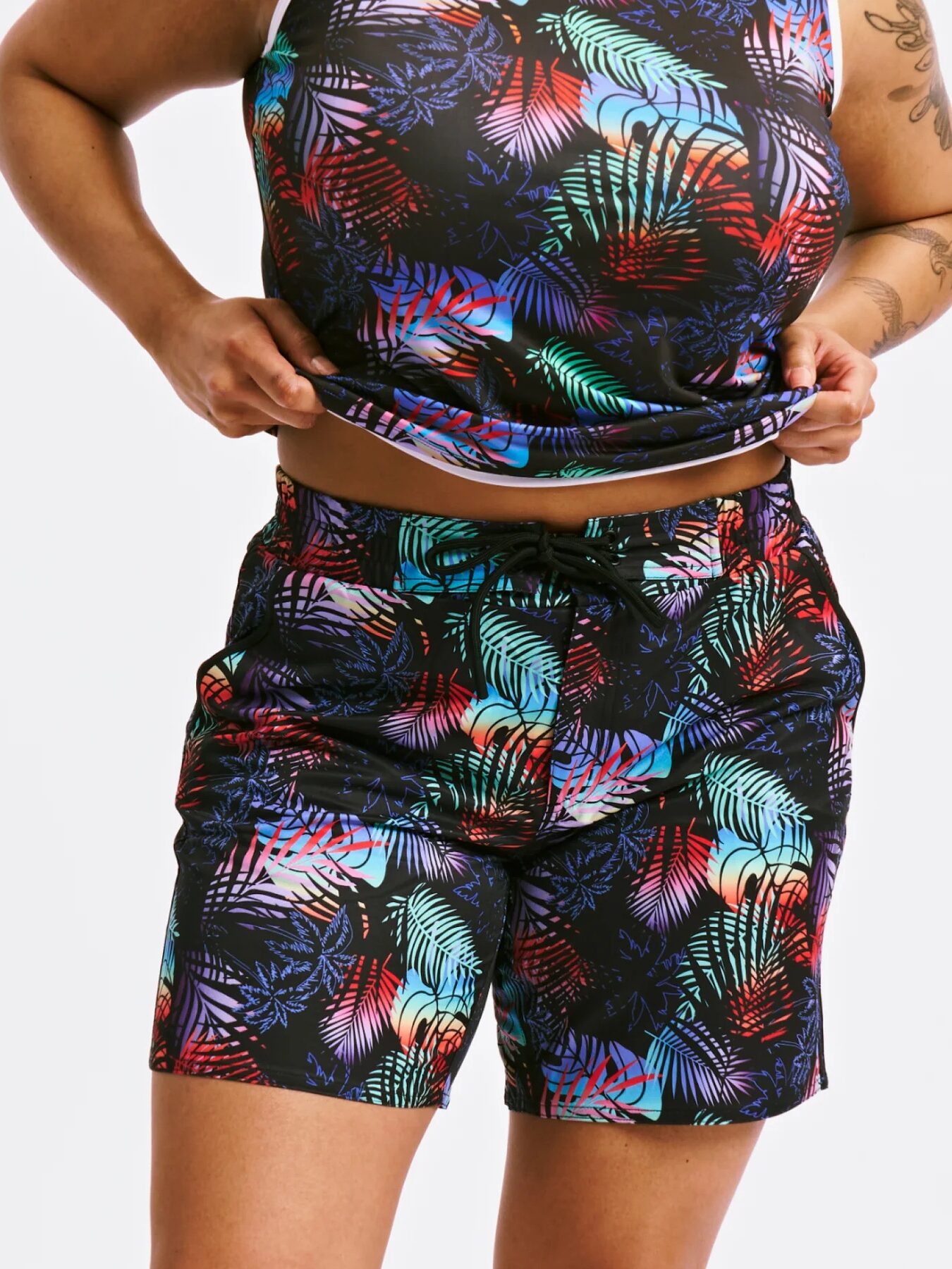 Person wearing a matching tank top and shorts set with a colorful leaf pattern on a black background, lifting the shirt slightly to show the shorts.