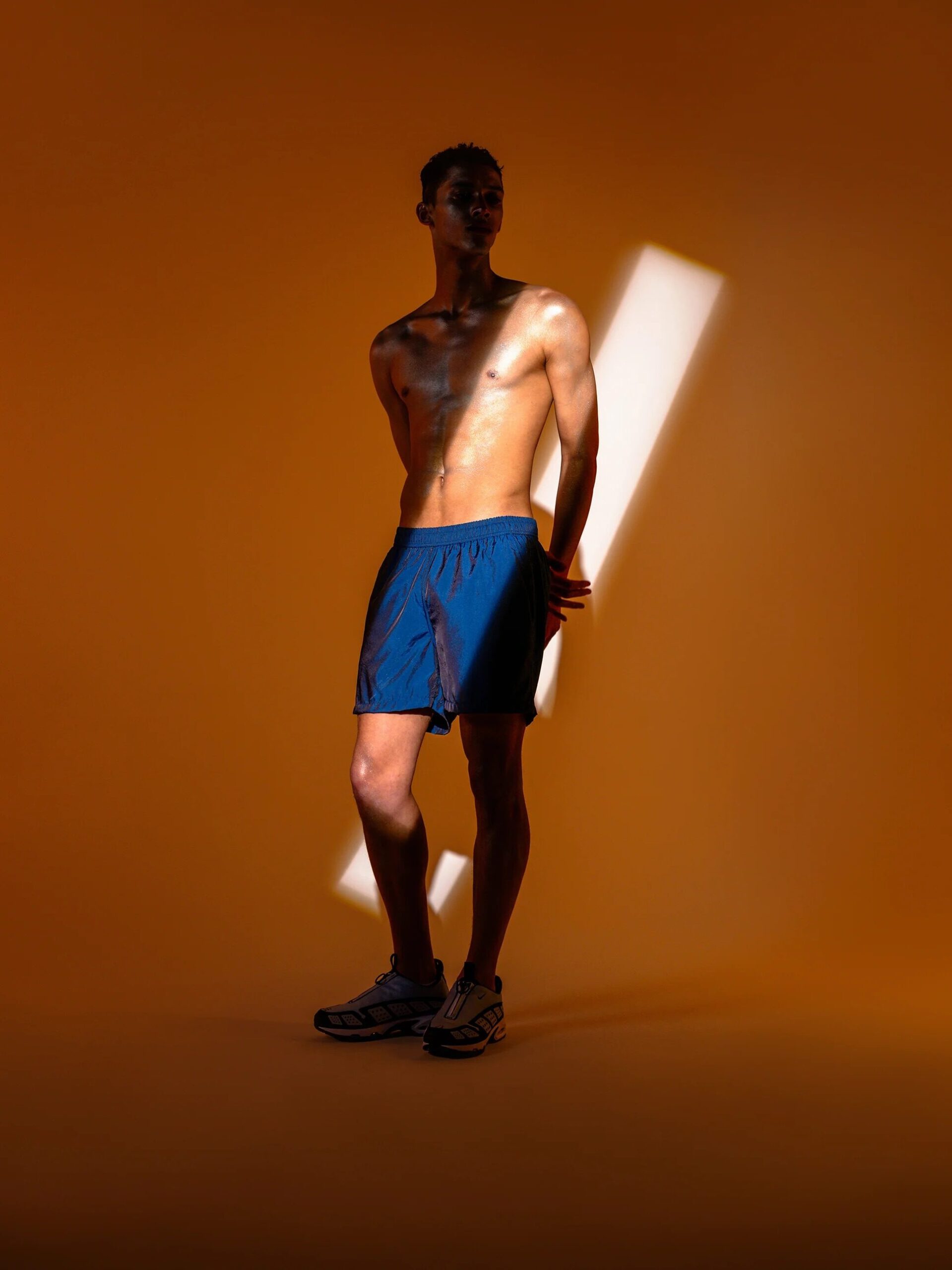 A shirtless man wearing blue shorts and sneakers stands against an orange background with a diagonal beam of light across his upper body.