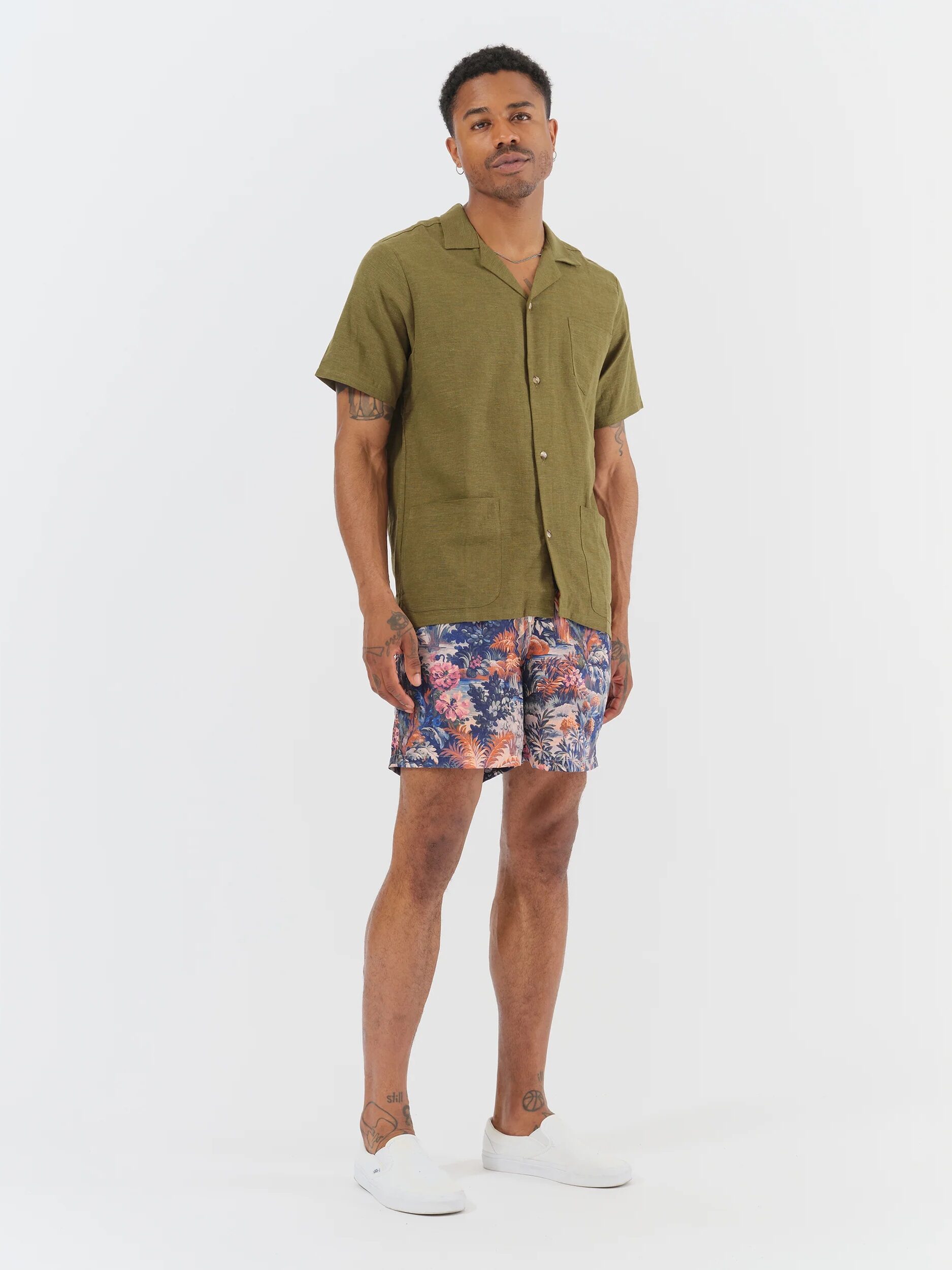 A man stands against a white background wearing an olive short-sleeve button-up shirt, colorful floral shorts, and white slip-on shoes.