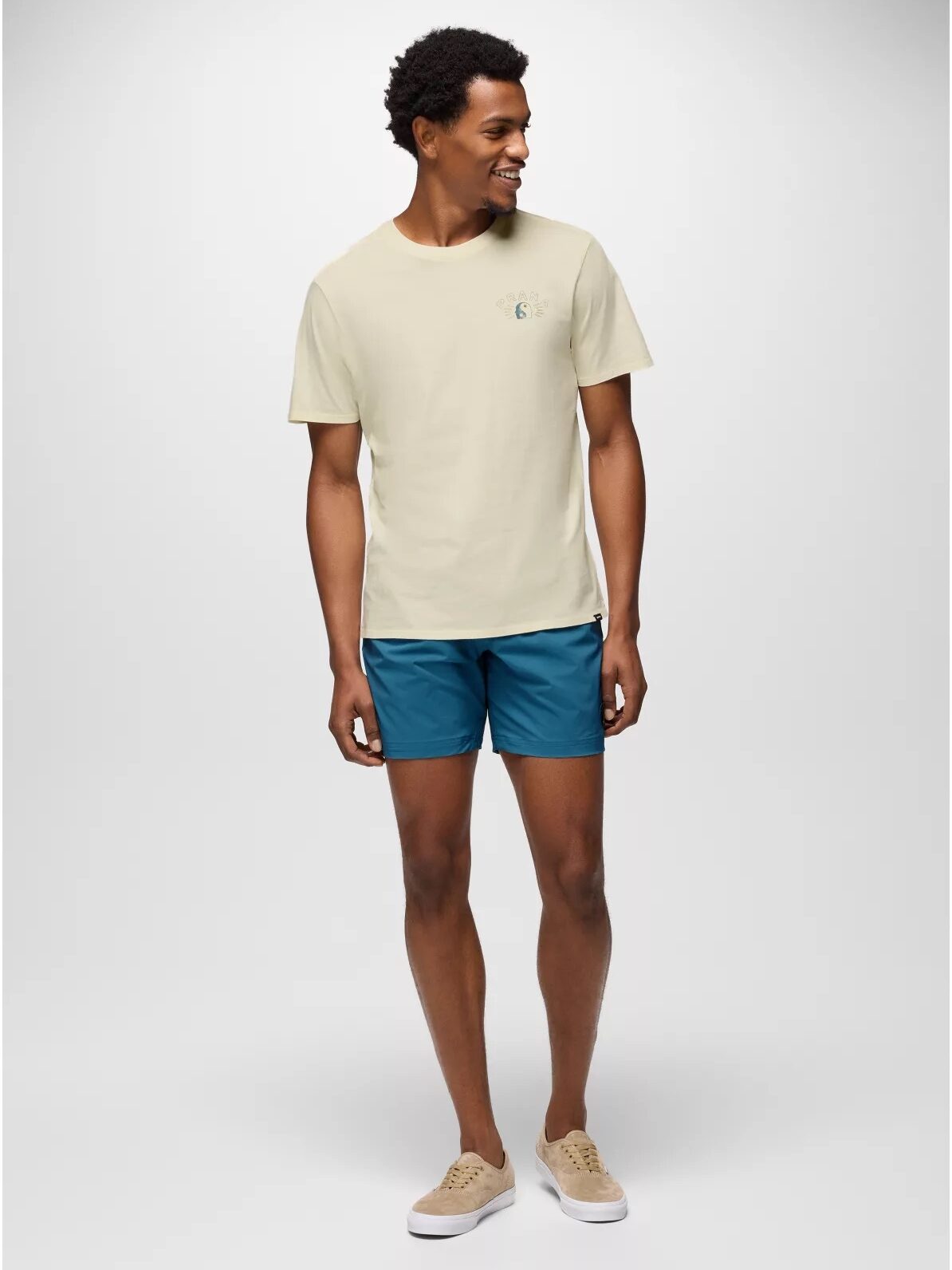 A man stands against a plain background wearing a light beige T-shirt, teal shorts, and beige sneakers. He is looking to his left and smiling.