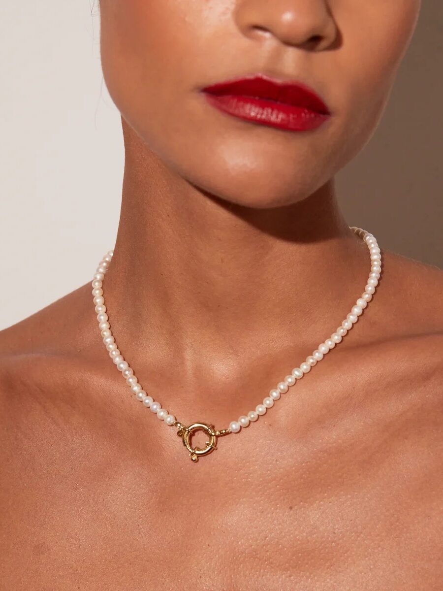 A woman wearing a strand of small pearls with a gold circular clasp necklace, red lipstick, and bare shoulders against a neutral background.