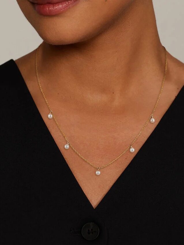 A woman wearing a black V-neck top and a delicate gold necklace with five evenly spaced dangling pearls.