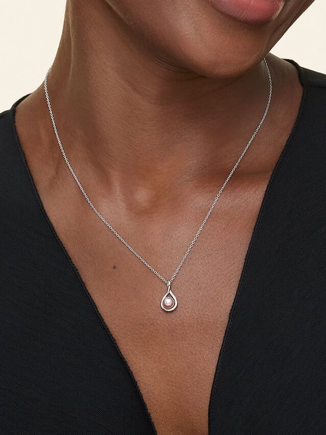 A person wearing a teardrop-shaped pendant necklace with a pearl on a thin chain, paired with a black V-neck top.