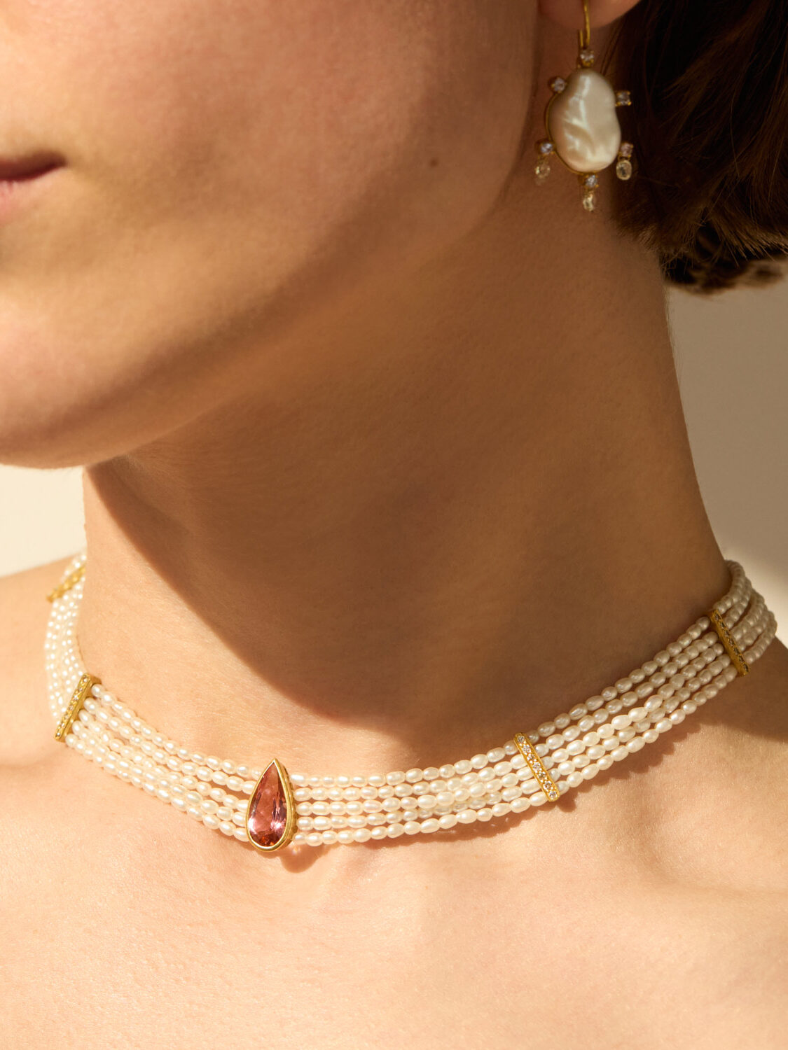 A person wearing a multi-strand pearl choker with a teardrop gemstone pendant and a matching pearl earring.
