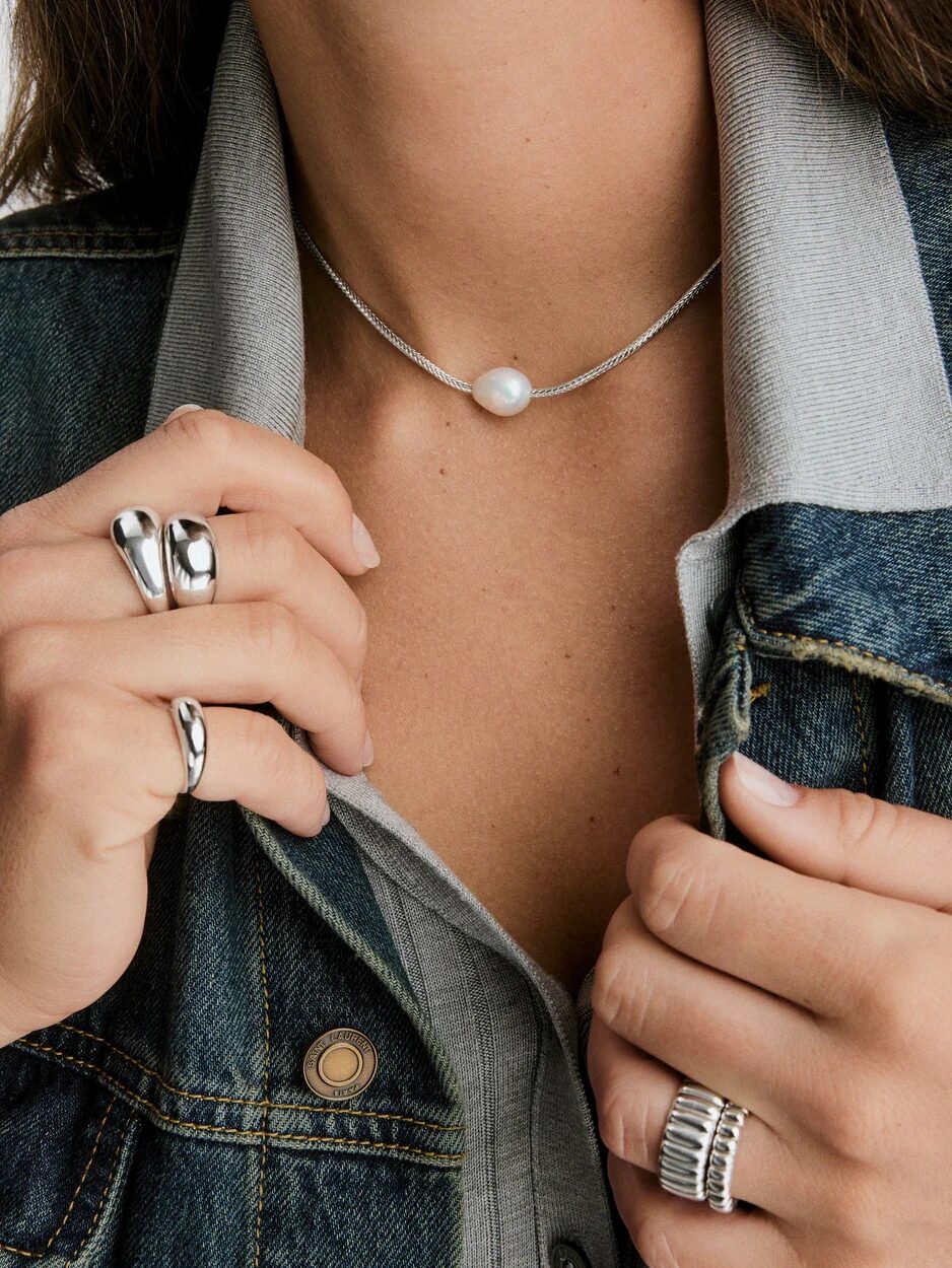 A person wearing a denim jacket, a collared shirt, a pearl necklace, and several silver rings on their fingers.