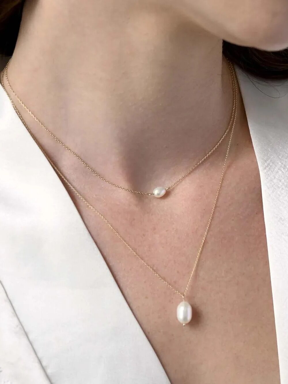A woman wearing a white satin robe displays two gold necklaces, each with a single white pearl pendant of different sizes.