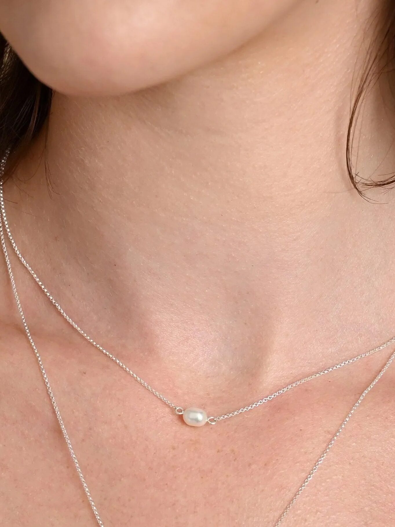 A close-up of a woman's neck wearing two delicate silver necklaces, one featuring a small pearl pendant in the center.