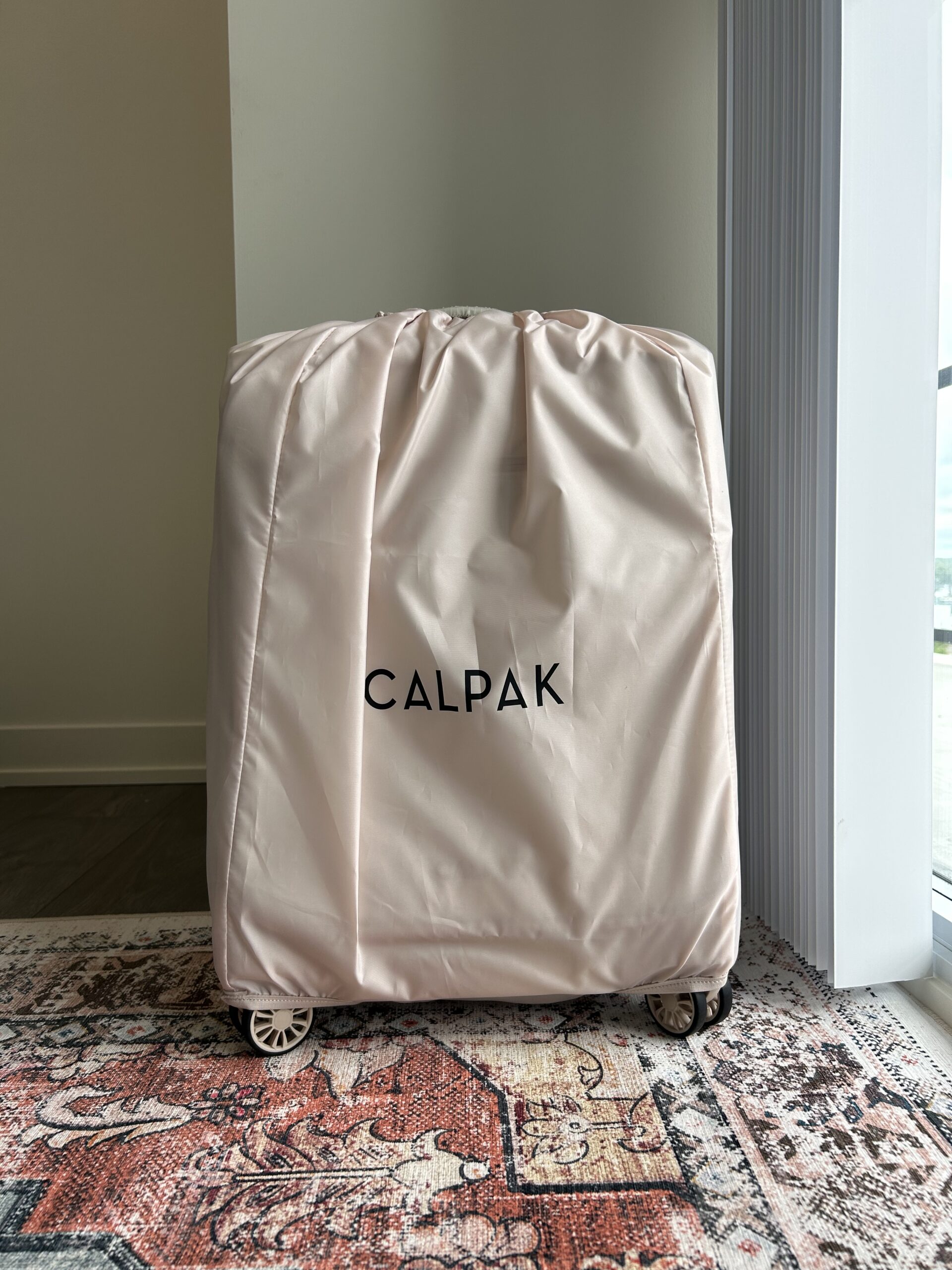 A light pink CALPAK suitcase cover is placed over a wheeled suitcase, positioned near a window on a patterned rug.