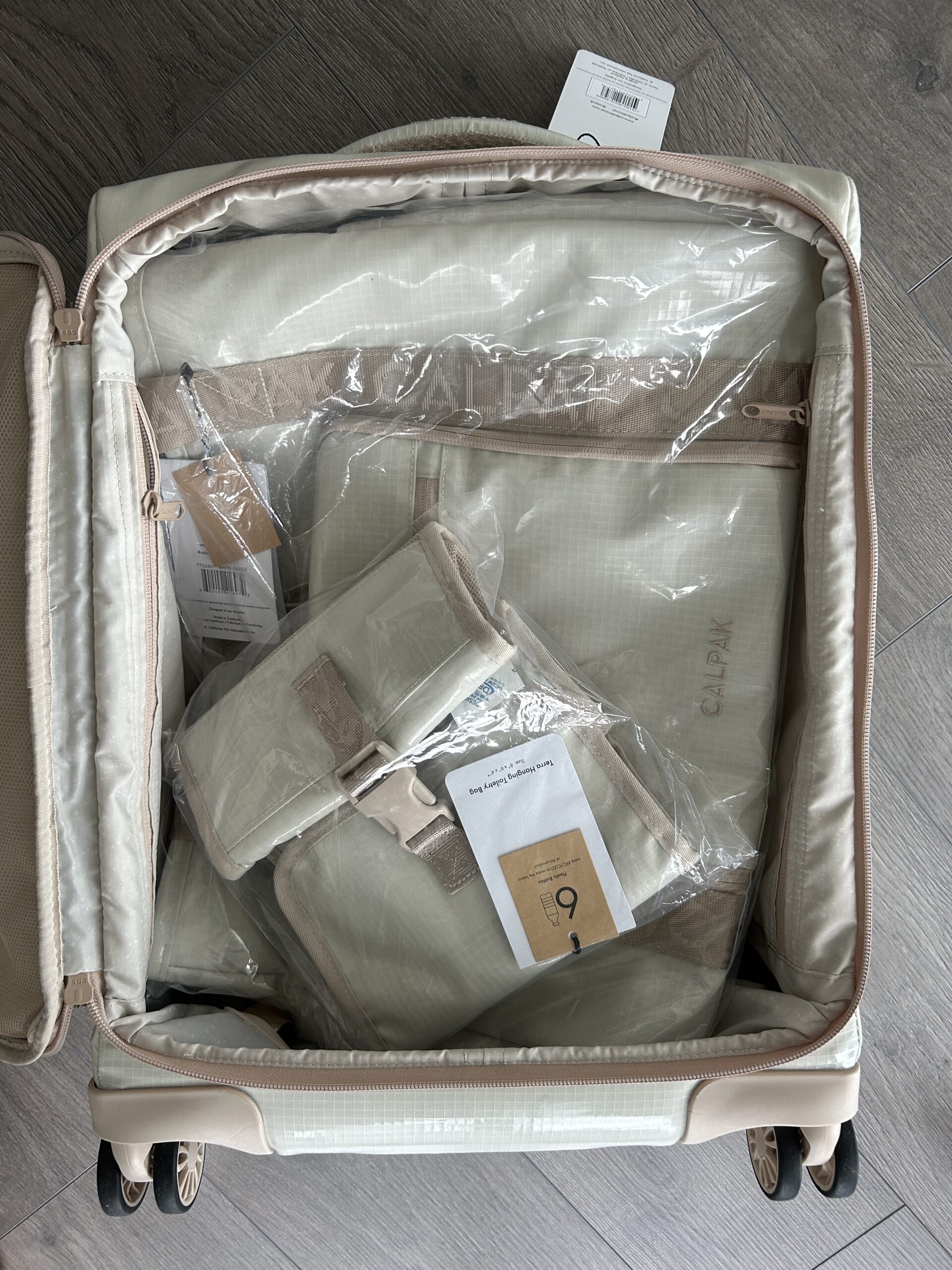 An open suitcase containing several beige packing cubes and bags in clear plastic packaging, placed on a gray wooden floor.