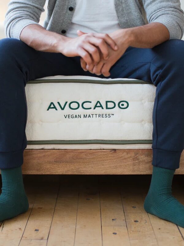 A person sits on the edge of a bed with "Avocado Vegan Mattress" branding, wearing blue pants and green socks.