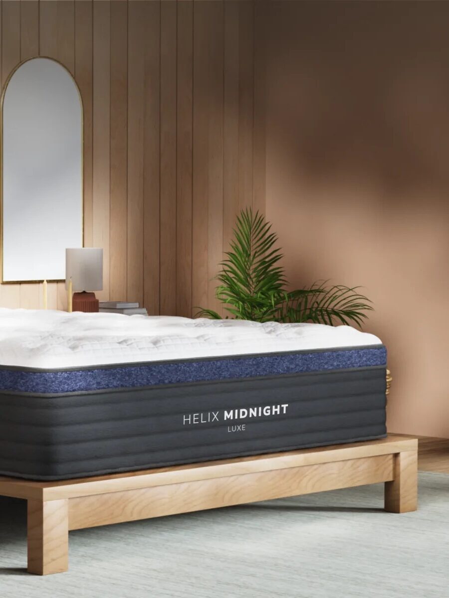 A bedroom with a wooden bed frame, a Helix Midnight Luxe mattress, white bedding, two bedside tables, mirrors, lamps, and potted plants against wood-paneled walls.