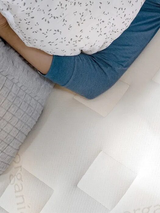 Person lying on a bed with a grey blanket, wearing a blue sleeve shirt, holding a white pillow with a subtle leaf pattern.