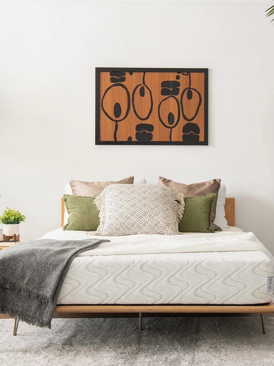 A modern bedroom with a neatly made bed, green and beige pillows, a side table, plants, a floor lamp, and abstract wall art above the bed. A "365 Night Sleep Trial" icon is at the bottom right.