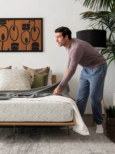 A man in casual clothes makes a neatly arranged bed in a modern, plant-filled bedroom with minimalist decor.