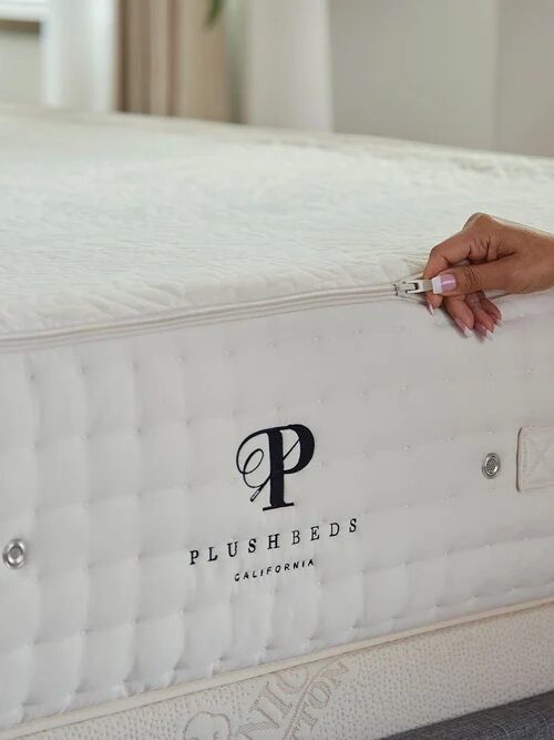 A hand unzips the cover of a white mattress with "PLUSHBEDS CALIFORNIA" and a "P" logo embroidered on the side.