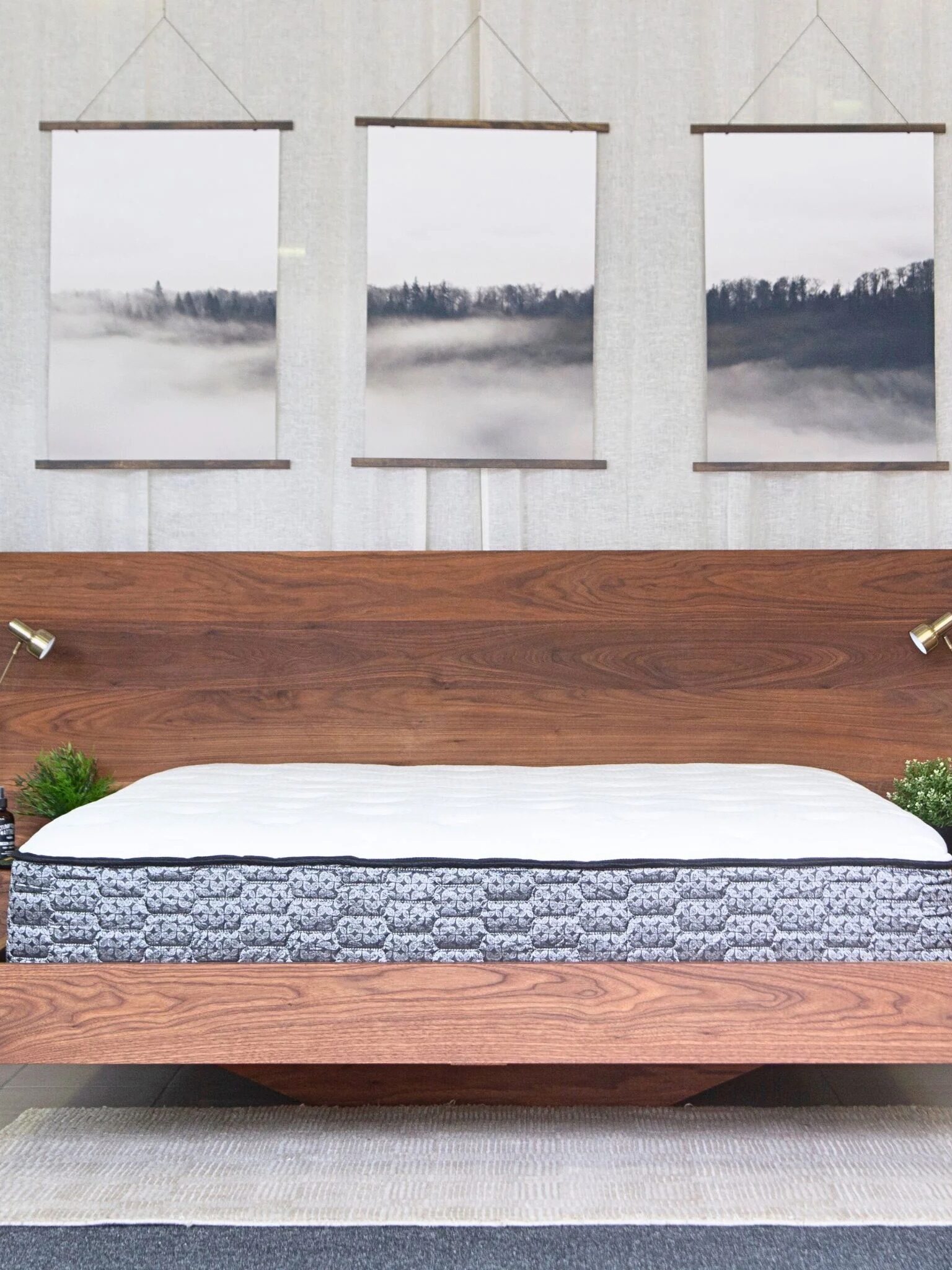 Modern wooden bed with a patterned mattress, flanked by two brass lamps and plants, with three landscape art prints hanging above against a white curtain backdrop.