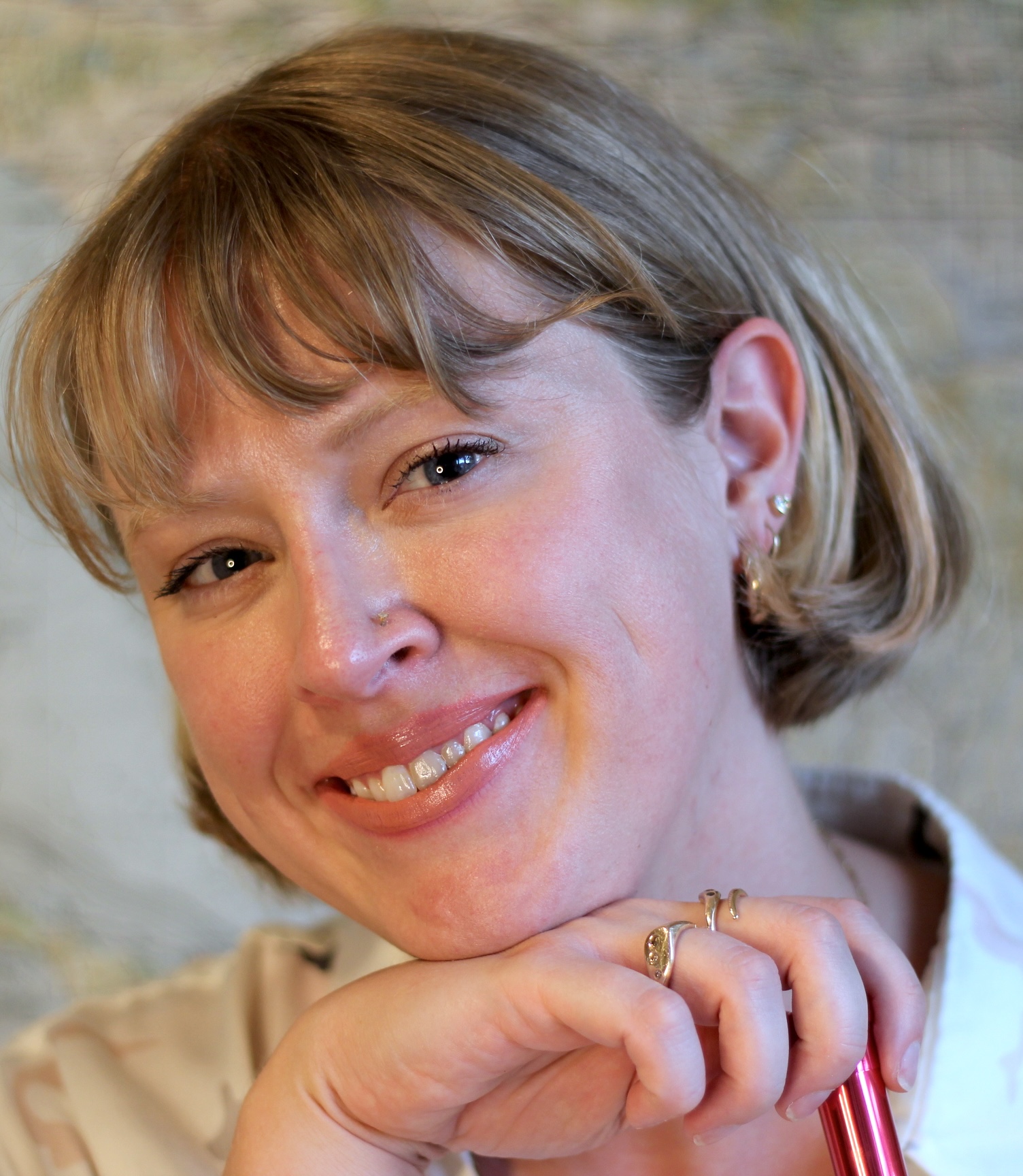 A woman with short blonde hair smiles at the camera, resting her chin on her hand and holding a red pen. She wears earrings and a light-colored top.