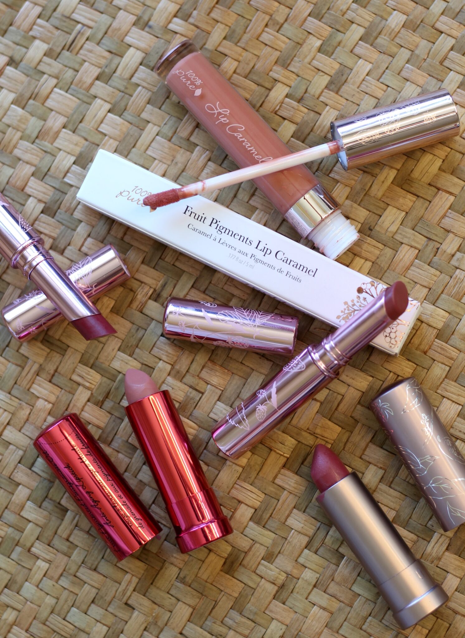 Several tubes of lipsticks and a lip gloss with their caps off are arranged on a woven surface, alongside a product box labeled "Fruit Pigments Lip Caramel.