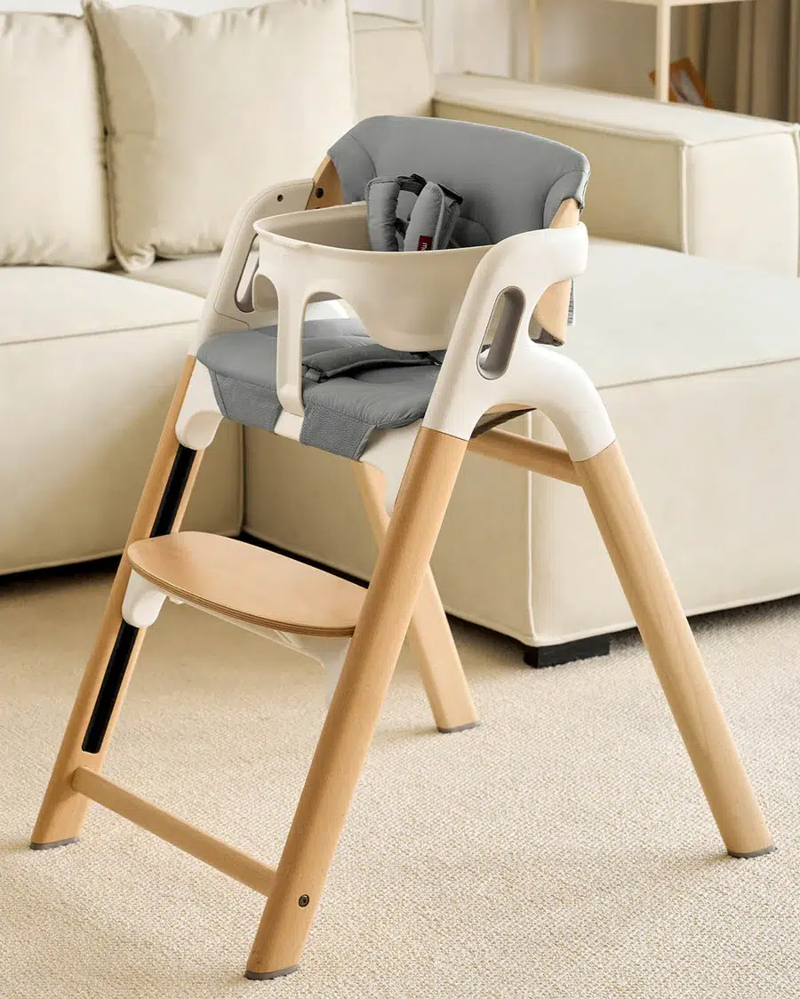 A modern wooden high chair with white and gray accents is placed on a carpeted floor in front of a beige sofa.