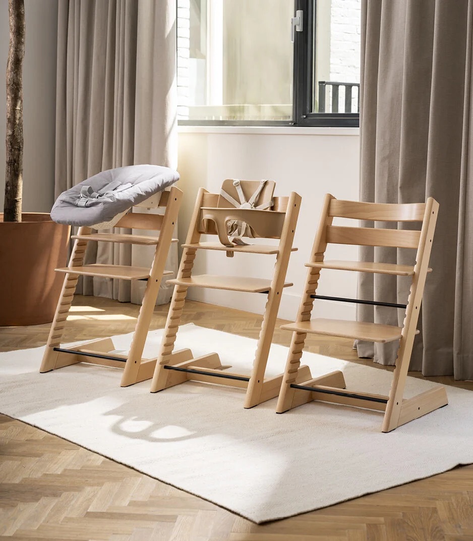 Three wooden adjustable high chairs are arranged side by side on a light rug in front of a window with beige curtains, with different seat and cushion configurations displayed.