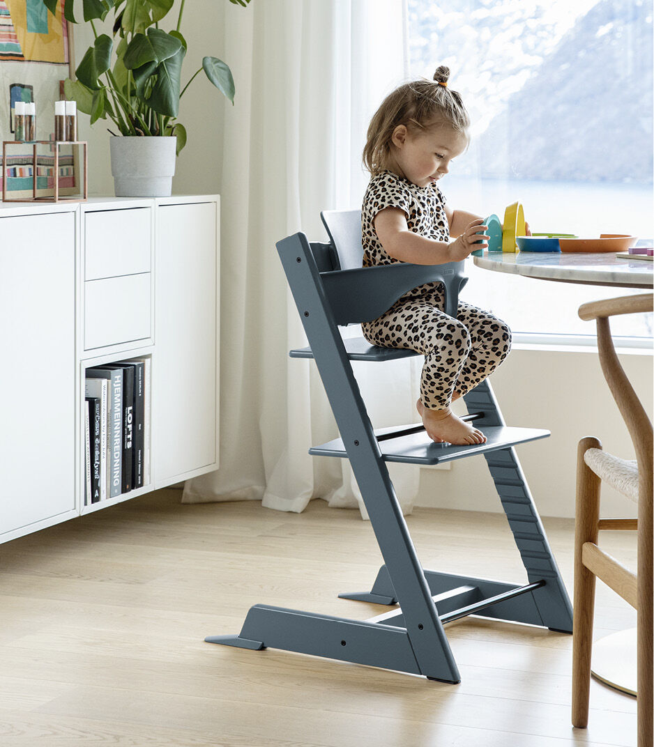 A young child sits on a modern high chair playing with toys at a table in a bright, minimalist room with large windows and light wood floors.