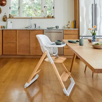 Modern wooden high chair with a white seat is placed near a dining table in a kitchen with wooden cabinets and light wood flooring.