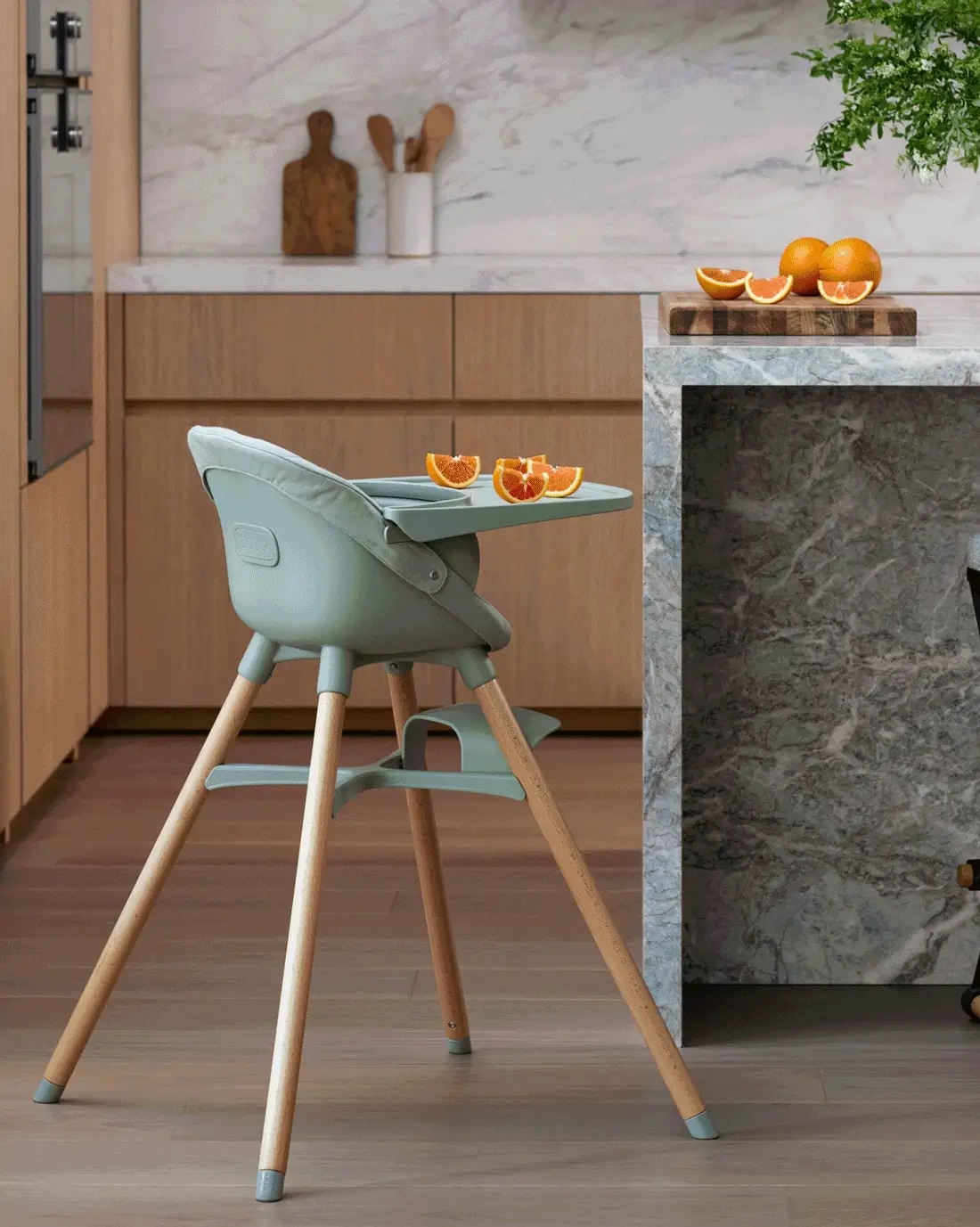A modern high chair with cut orange slices on its tray is positioned next to a kitchen island with more sliced oranges and a leafy green plant.