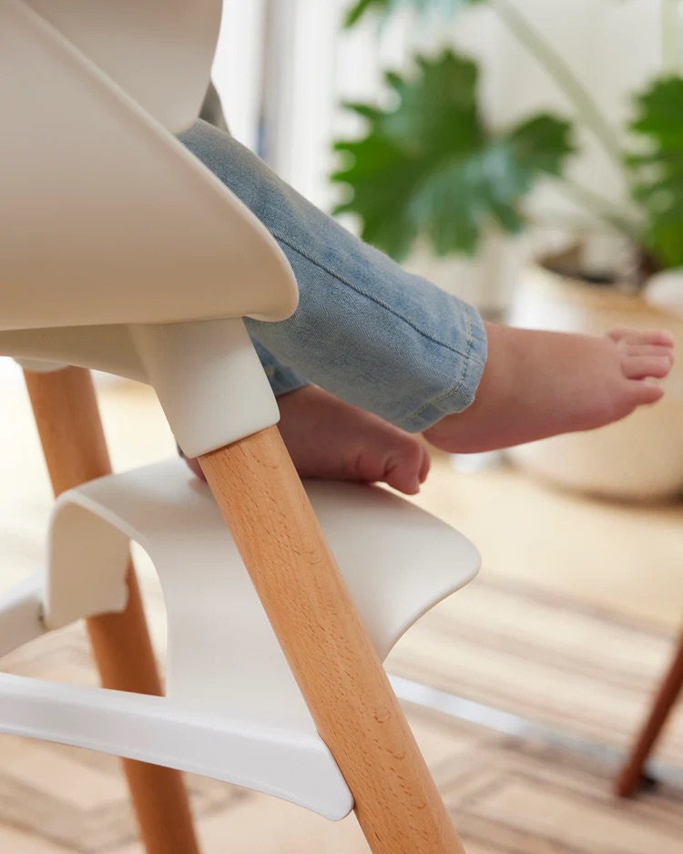 A child's bare feet rest on the footrest of a white and wooden high chair, with a leafy plant in the background.