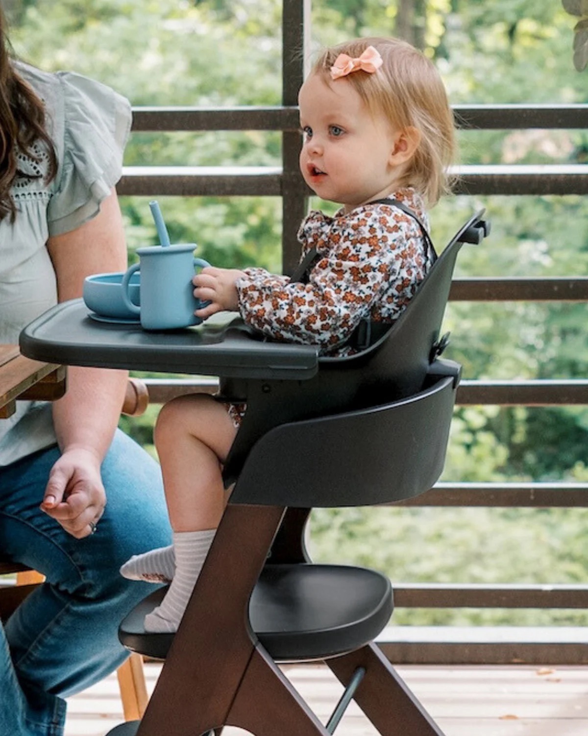 A toddler sits in a high chair wearing a floral outfit with a bow in her hair, holding blue child-friendly dishware, beside an adult on a wooden patio.