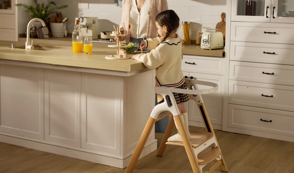 A young girl stands on a wooden step stool at a kitchen island while an adult prepares food beside her; two glasses of orange juice are on the counter.