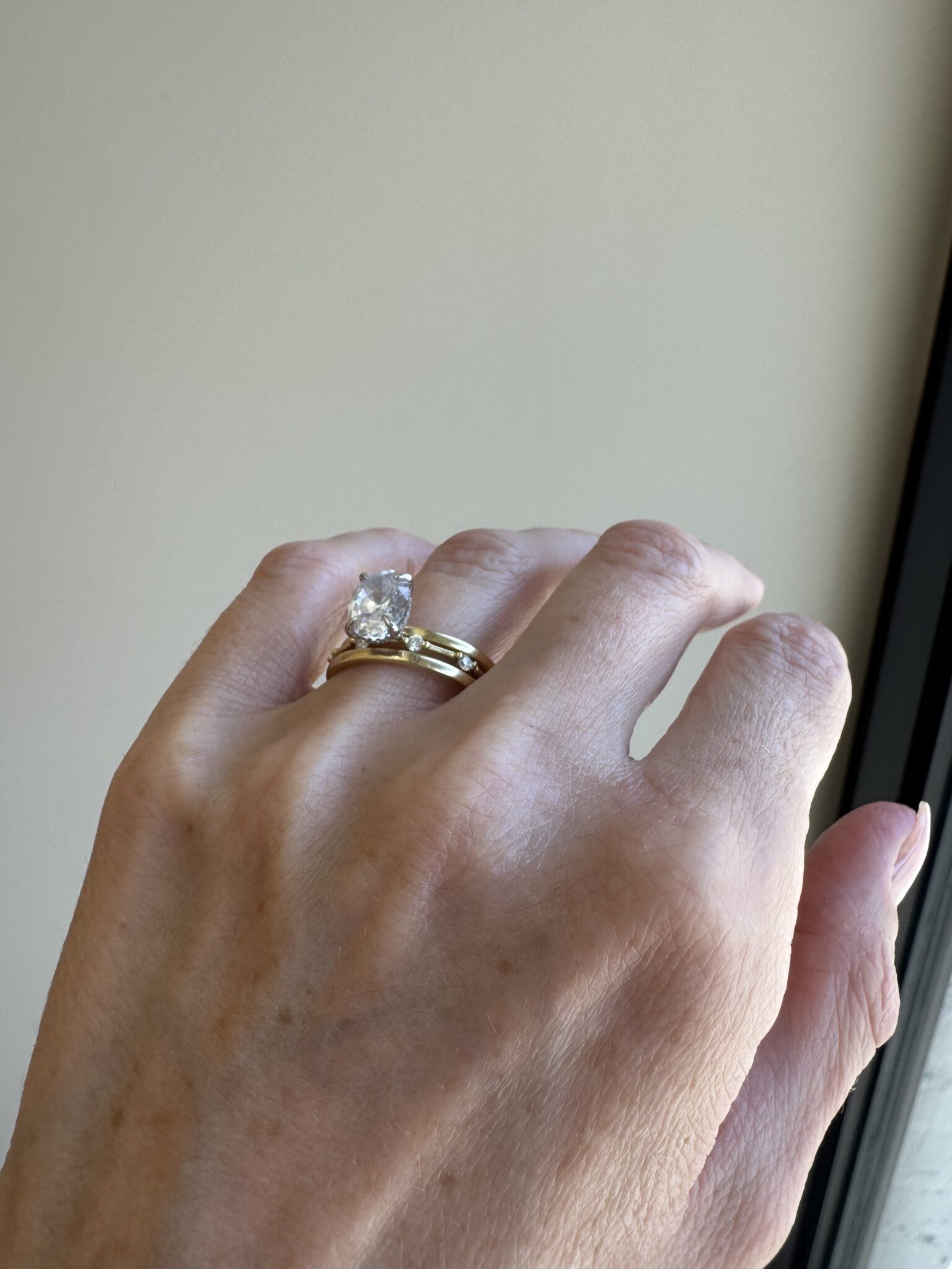 A hand wearing a gold wedding band and a large diamond engagement ring, shown against a plain background near a window.