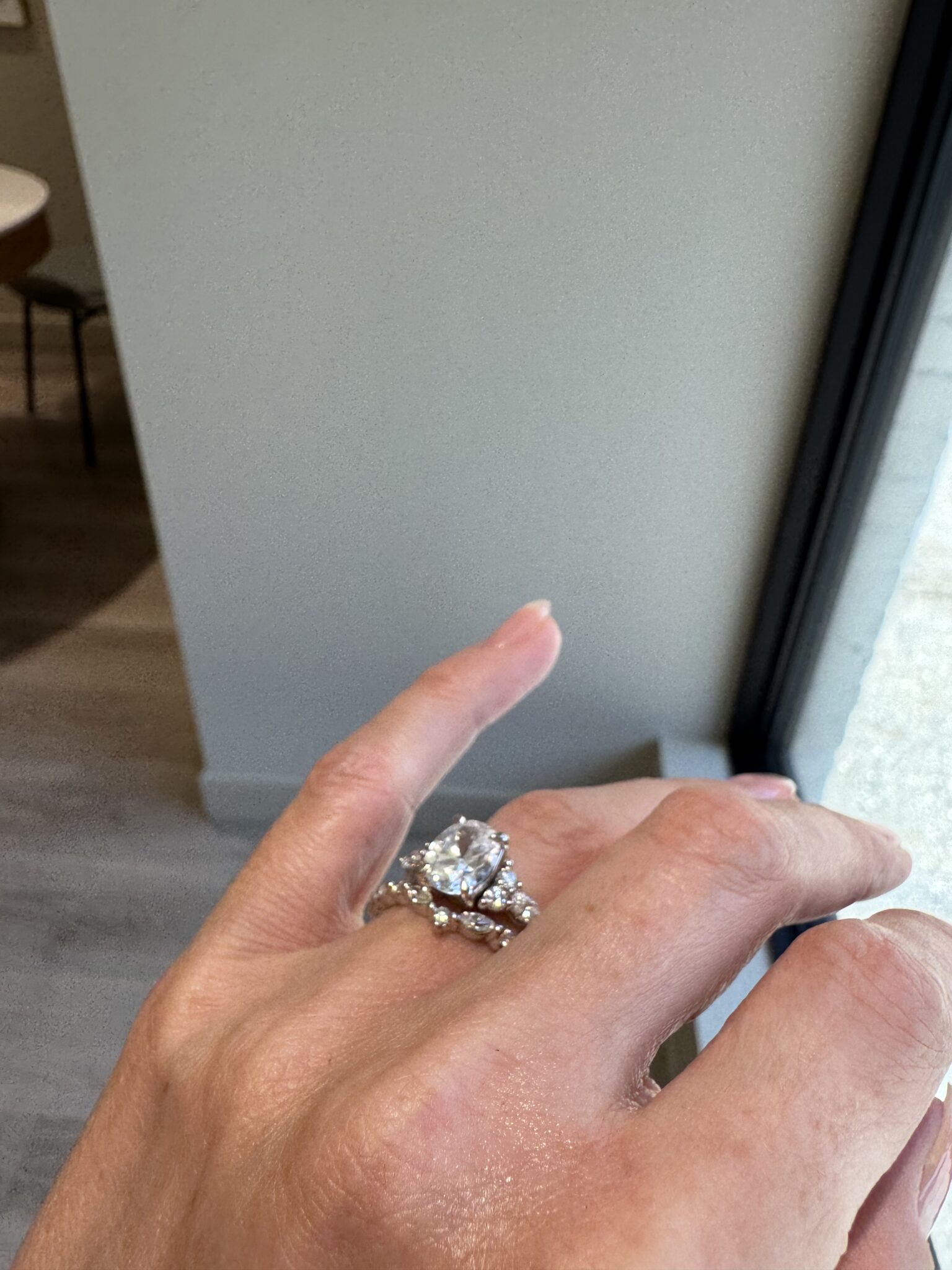 A hand with a large, round-cut diamond ring and an additional band is held up near a window, with part of a room and floor visible in the background.