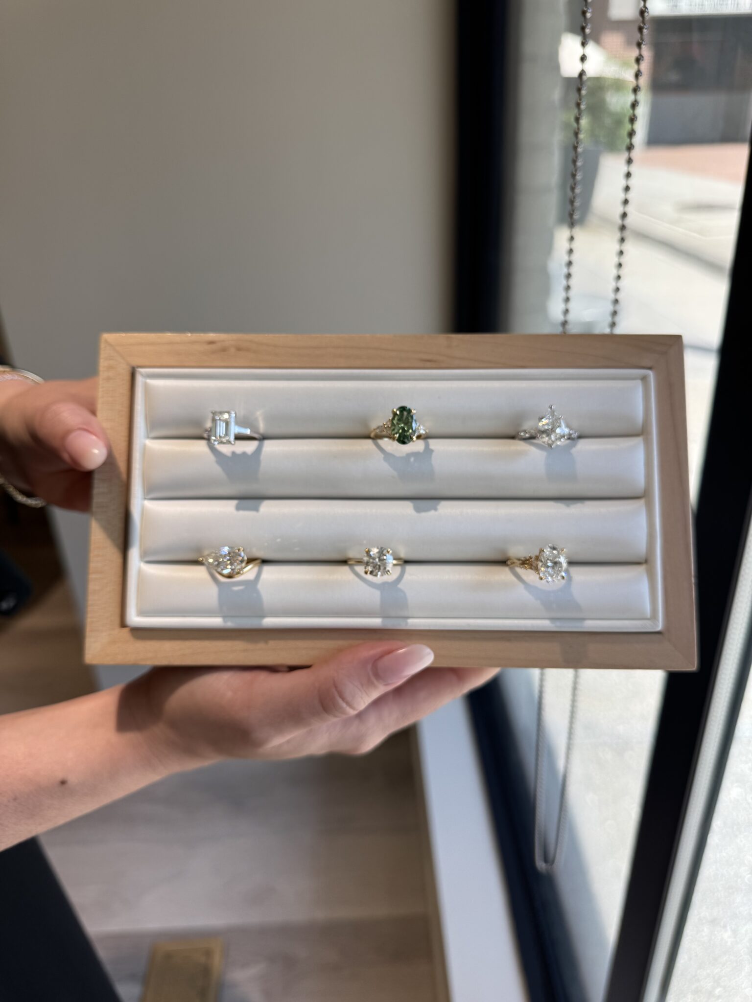 A person holds a tray with six different gold rings, each with a unique gemstone or diamond, displayed in a jewelry store near a window.