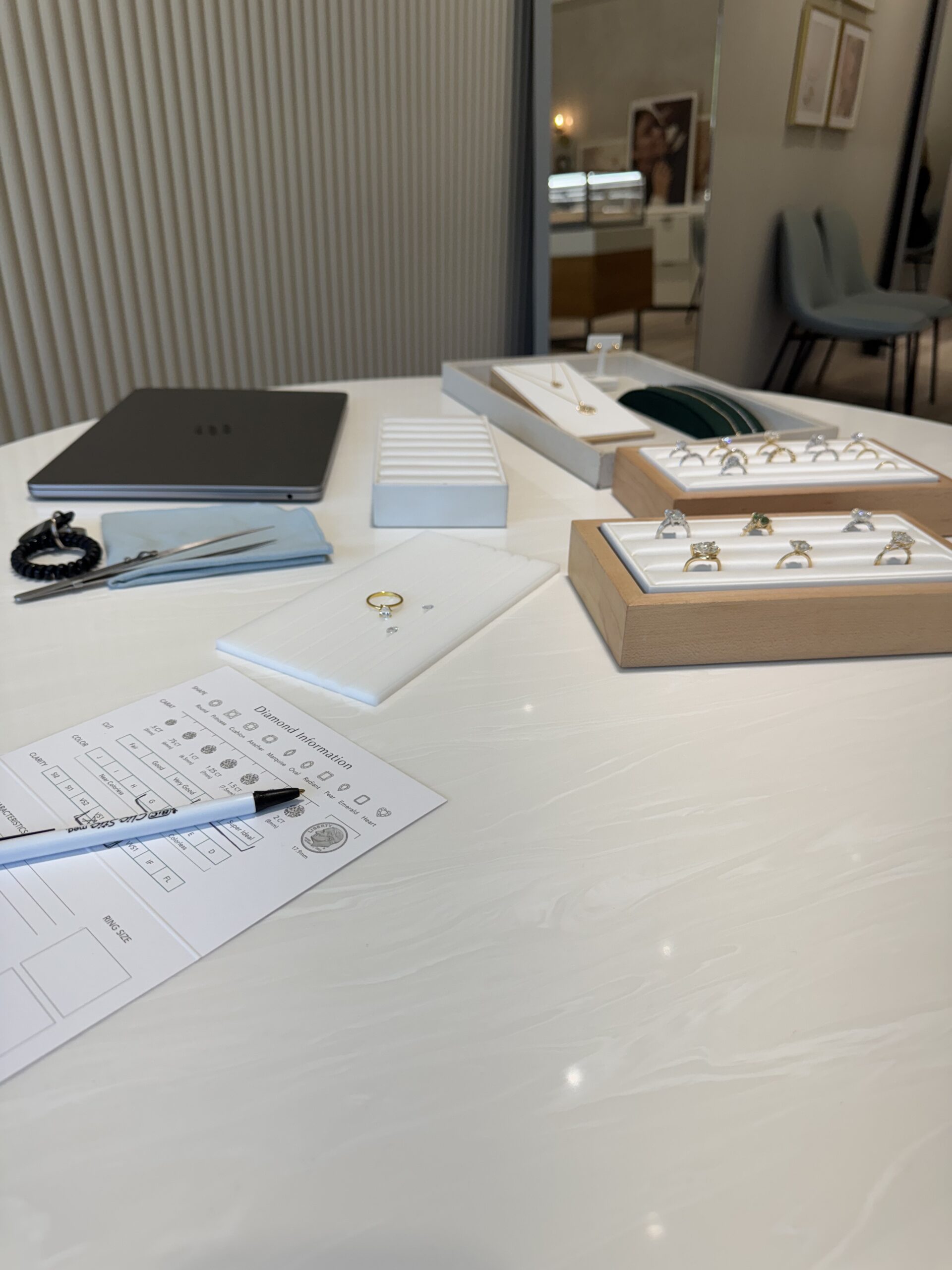 A jewelry store counter displays various rings, a laptop, a pen, a ring sizing chart, and a notepad on a white surface.